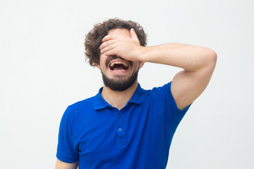 Fototapeta na wymiar Hysterical guy covering eyes and face with hand and shouting. Handsome bearded young man in blue casual t-shirt posing isolated over white background. Annoyance concept