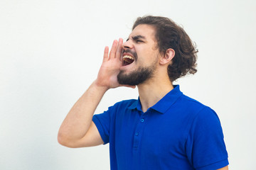 Excited guy sharing important news, shouting loud with hand at open mouth. Handsome bearded young man in blue casual t-shirt posing isolated over white background. Advertising concept