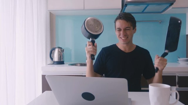 crazy funny man is dancing with a frying pan and a pan in his hands while sitting at a laptop in the kitchen celebrating a victory or achievement.