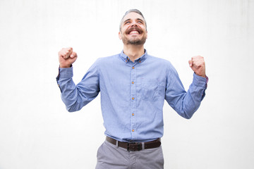Fototapeta premium Excited overjoyed man making victory or winner gesture, looking up, smiling. Grey haired young man in blue casual shirt posing isolated over white background. Luck or fortune concept