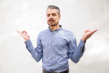 Tranquil focused man making zen gesture with hand and fingers. Grey haired young man in blue casual shirt posing isolated over white background. Meditation concept