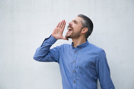 Excited man announcing news and shouting loud with hand at mouth. Grey haired young man in blue casual shirt posing isolated over white background. Announcement concept