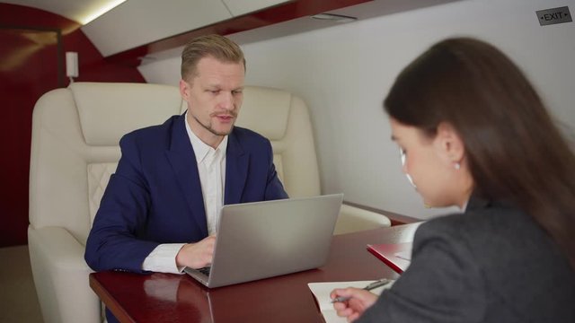 Zoom out medium shot of businessman with laptop giving directions to young female secretary making notes in notebook sitting at desk during flight on private jet