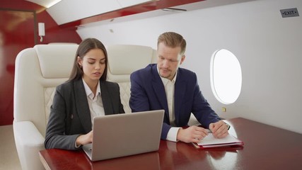 Middle aged businessman and young businesswoman sitting at desk during flight on private jet and discussing data on laptop computer - Powered by Adobe