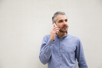 Pensive man speaking on cell, looking away into distance. Grey haired young man in blue casual shirt posing isolated over white background. Phone conversation concept