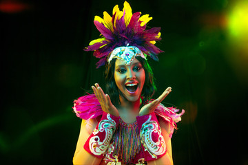 Beautiful young woman in carnival mask and stylish masquerade costume with feathers in colorful lights and glow on black background. Christmas, New Year, celebration. Festive time, dance, party.