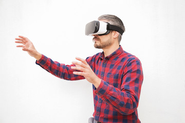 Man in VR headset touching air. Serious bearded man in checkered shirt moving with hands and using VR headset on grey background. Technology concept