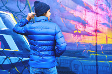 back view of young attractive woman wearing in blue down jacket and knitted hat posing against wall painted with graffiti. Urban clothing style, urban background.