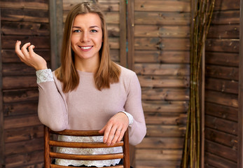 Loft style concept. Portrait of a pretty blonde girl in a pink sweater on a wooden background in the home interior. Cute looks and smiles at the camera, sitting on a chair straight