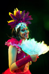 Beautiful young woman in carnival mask and stylish masquerade costume with feathers fan in colorful lights and glow on black background. Christmas, New Year, celebration. Festive time, dance, party.