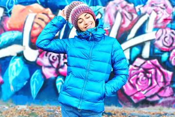 Portrait of young smiling woman wearing in blue down jacket and knitted warm hat posing against wall painted with graffiti. Urban clothing style, urban background.