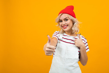 Beautiful young woman with short blonde curly hair and bright makeup in white overalls and red hat likes something and smiles, portrait isolated on orange background