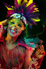 Beautiful young woman in carnival mask and stylish masquerade costume with feathers in colorful lights and glow on black background. Christmas, New Year, celebration. Festive time, dance, party.