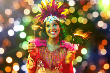 Beautiful young woman in carnival mask and stylish masquerade costume with feathers and sparklers in colorful bokeh on black background. Christmas, New Year, celebration. Festive time, dance, party.