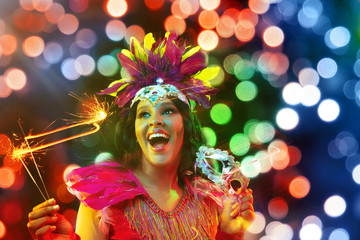 Beautiful young woman in carnival mask and stylish masquerade costume with feathers and sparklers in colorful bokeh on black background. Christmas, New Year, celebration. Festive time, dance, party.