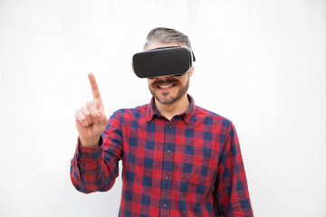Naklejka premium Content man in VR headset pointing with finger. Front view of cheerful bearded man in checkered shirt using virtual reality headset on grey background. Technology concept