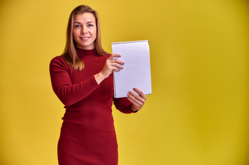 Fototapeta na wymiar Portrait of a pretty blonde girl with a beautiful smile and excellent teeth in a burgundy dress with a folder in her hands on a yellow background. Cute looks at the camera, standing straight.