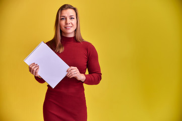 Portrait of a pretty blonde girl with a beautiful smile and excellent teeth in a burgundy dress with a folder in her hands on a yellow background. Cute looks at the camera, standing straight.