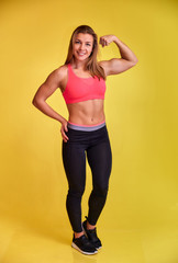 Fototapeta na wymiar Vertical portrait of a pretty fitness blonde girl with a beautiful smile, excellent teeth and a bodybuilder figure in a tracksuit on a yellow background. Cute looks at the camera, standing straight.