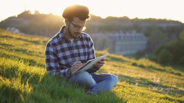 Handsome Young Artist Sketching and Drawing Landscape While Sitting in Grass During Colorful Sunset