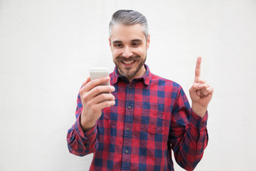 Content man with smartphone pointing up with finger. Cheerful bearded man gesturing with finger and using mobile phone on grey background. Idea concept