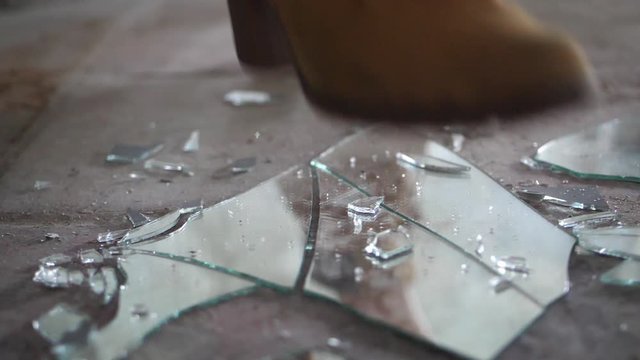 A close up shot a woman with a pair of suede brown boots, kicking down on a piece of mirror, cracking and destroying the glass into small pieces.