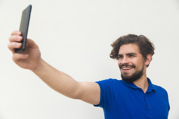 Cheerful guy taking selfie on mobile phone. Handsome bearded young man in blue casual t-shirt...