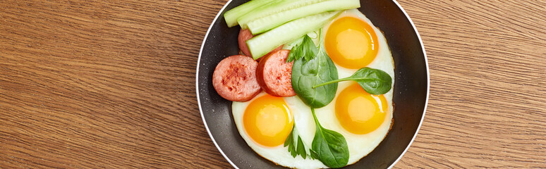 top view of fried eggs with spinach leaves, cucumber and sausage in frying pan on wooden table, panoramic shot