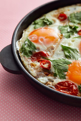 close up view of fried eggs with parsley, tomatoes and chili pepper in frying pan on pink background