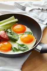 selective focus of fried eggs in frying pan with spinach, cucumber and sausage at wooden table with cutlery on napkin