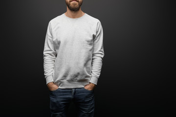 partial view of bearded man in blank basic grey sweatshirt with hands in pockets isolated on black