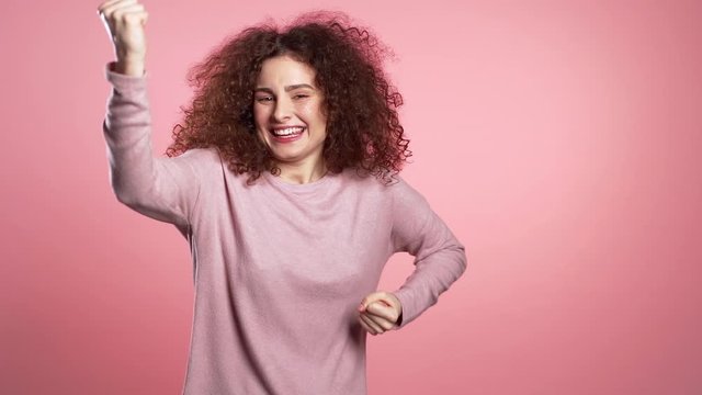 Girl with curly hair and extraordinary makeup shows yes gesture of victory, she achieved result, goals. Surprised excited happy woman on pink background
