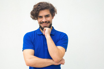 Fototapeta na wymiar Positive happy guy leaning chin on hand, looking at camera, smiling. Handsome bearded young man in blue casual t-shirt posing isolated over white background. Male portrait concept