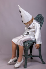 Cute child dressed like Pierrot, sad clown. Mascarade costume for holidays. Studio shot on gray...