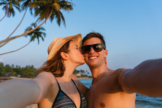 young happy couple taking a selfie in a tropical beach during sunset, girlfriend kissing boyfriend