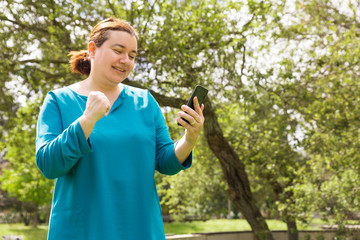 Happy lucky woman with mobile phone reading message, getting good news while walking in park. Middle aged woman in casual posing outdoors. Success concept