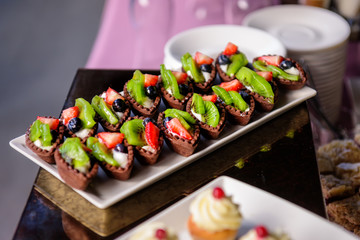 Delicious sweet tartlets made with chocolate dough with fresh fruit and berries close-up. Small cakes with strawberries, kiwi, grapes. Banquet service at wedding, anniversary.