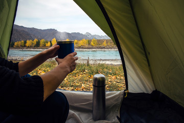 View from the tent on river and mountains. Traveler woman sitting in a tent holds a thermo cup in her hand, close up. Traveling and spending time in tent while drinking coffee in the morning.