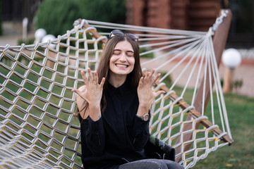 Happy woman in dark shirt, jeans and sunglasses showing a ring and relaxing in hammock in forest at...