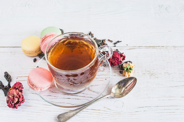 Tea time concept. Glass cup of black tea, french macaroons and dry rose buds on the rustic white paint wooden backgorund