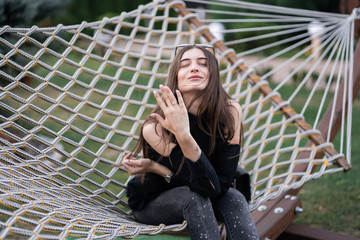 Happy woman in dark shirt, jeans and sunglasses showing a ring and relaxing in hammock in forest at...