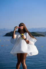 Young beautiful woman in white dress and sunglasses blowing soap bubbles on pier with seaview background. The concept of joy, ease and freedom during the vacation. The girl is enjoying the rest.