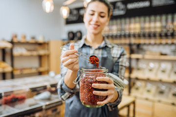 Young Mixed Race Woman Owner Selling Superfoods in Zero Waste Shop. Lots of Healthy Food in Glass Bottles on Stand in Grocery Store. No plastic Conscious Minimalism Vegan Lifestyle Concept. Copy space