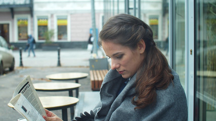 Side view, young woman in a grey blanket and reads a newspaper