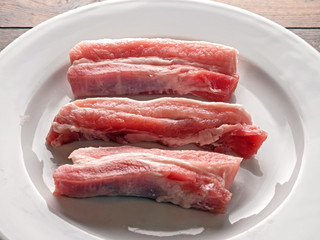 Uncooked raw pork belly slices on a white plate and wooden table.