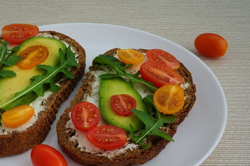 Sandwich with fresh vegetables arugula, avocado and tomatoes on bread with cheese in a white plate on a beige background