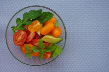 Salad with fresh vegetables arugula, avocado and tomatoes in a glass plate on a gray background with place for copy space, flat lay.
