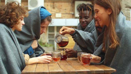 Group of friends in blankets sit outside the cafe and pour tea in cups