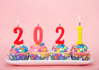 White cupcakes with rainbow colored frosting and brightly colored star candies on a rectangular plate pink background with 2021 candles burning. Happy New Year theme.