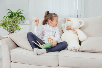 Nice little girl sitting on the sofa with teddy-bear near her, reading fairy-tale, pointing her finger up, saying something important to her soft friend.
